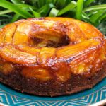 Bolo de banana caramelado inteiro com bananas caramelizadas na parte de cima, sobre prato turquesa e base de cobre