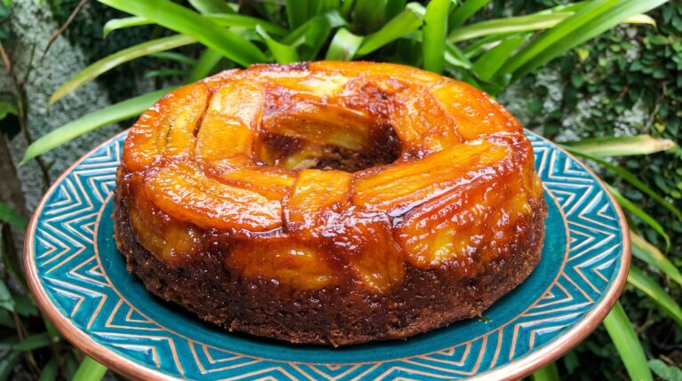 Bolo de banana caramelado inteiro com bananas caramelizadas na parte de cima, sobre prato turquesa e base de cobre